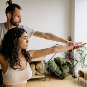 A yoga instructor helps a woman with her Warrior Pose in a serene indoor studio.