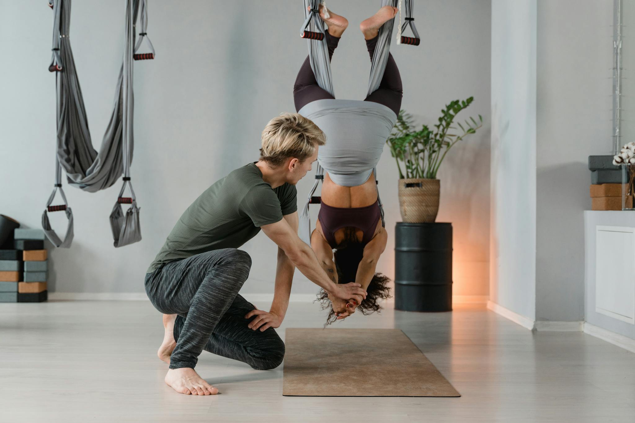 Aerial yoga class with an instructor aiding a student on a suspended hammock indoors.